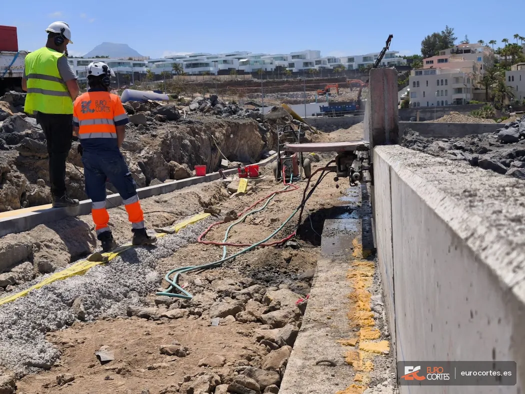 Sierra mural cortando un muro de hormigón junto a la costa mientras dos operarios supervisan el trabajo.